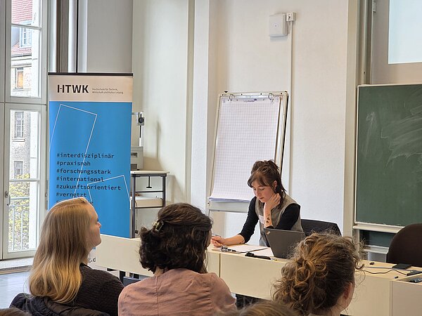 Keynote von Pauline Seuß Pauline Seuß sitzt an einem Tisch und schaut auf ihre Notizen. Vor ihr sitzen drei Personen und hören zu.