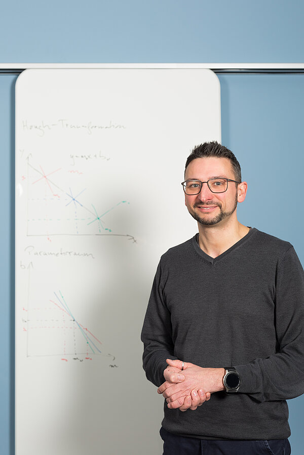 Dr. Mirco Fuchs Dr. Mirco Fuchs steht vor einem Whiteboard, auf dem Skizzen für Algorithmen zu sehen sind