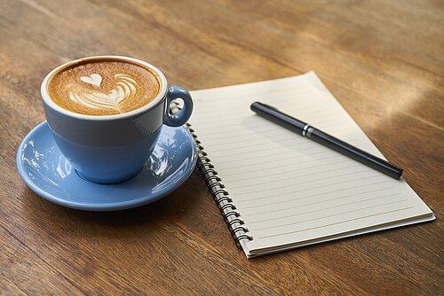 Chat Up! - Mit Kaffee & Unternehmen aus Sachsen. Eine Tasse mit Kaffee steht auf einem Holztisch, daneben liegt ein Notizzettel und ein Schreibtisch.