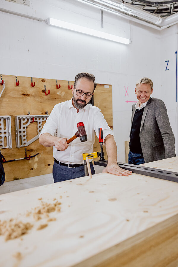 Sebastian Gemkow hämmert eigenhändig einen Holzdübel in eine Holzwandkonstruktion, neben ihm steht Prof. Alexander Stahr