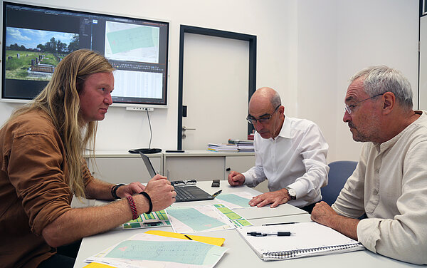 Besprechung Geotechnik HTWK-Mitarbeiter Felix Oertel, Prof. Ralf Thiele und Prof. Ronald Scherzer-Heidenberger besprechen die Messergebnisse der geotechnischen Untersuchungen am Tisch sitzend. Im Hintergrund sind auf einem großen Bildschirm Pläne und Fotos zu sehen.