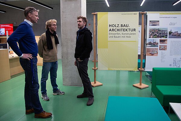 Im Foyer in der Hochschulbibliothek stehen Ausstellungstafeln, Personen besichtigen diese