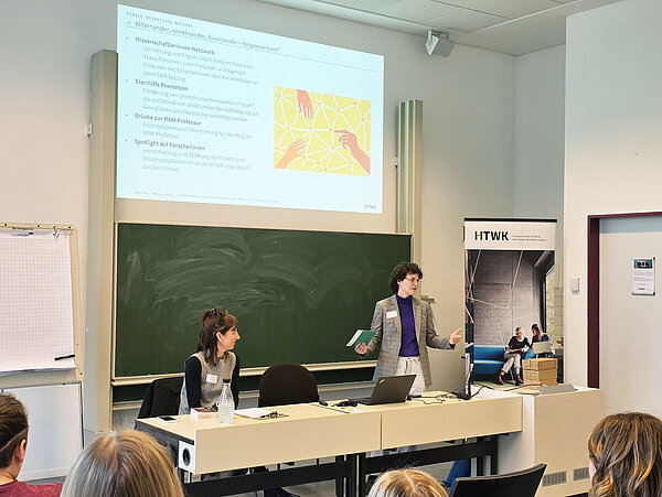 Keynote von Pauline Seuß Pauline Seuß und eine Moderatorin befinden sich vor einer Tafel. Die Moderatorin spricht und stellt die Veranstaltung vor.